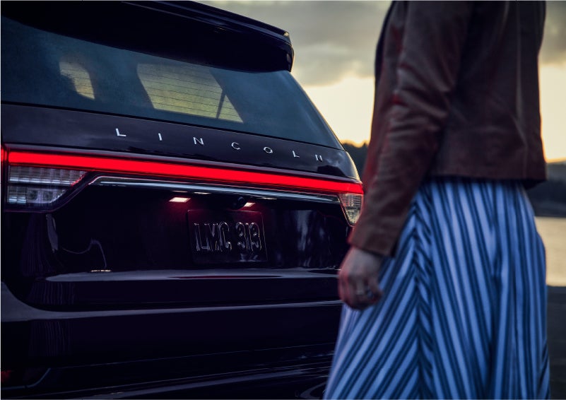 A person is shown near the rear of a 2023 Lincoln Aviator® SUV as the Lincoln Embrace illuminates the rear lights | Mathews Lincoln in Marion OH