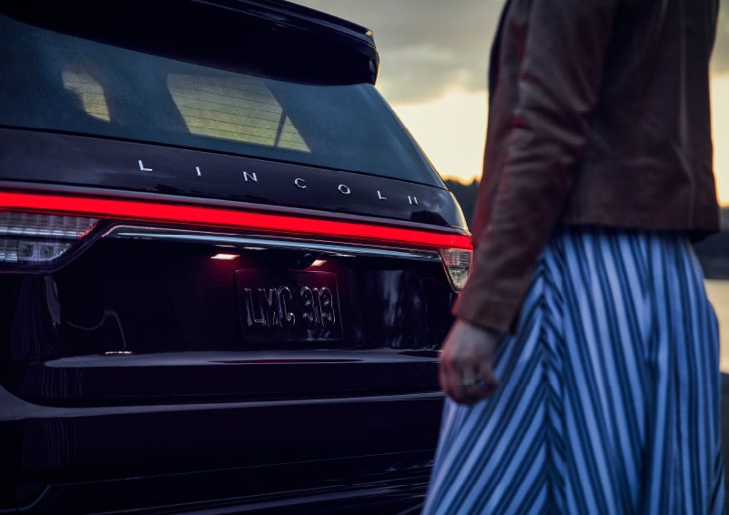 A person is shown near the rear of a 2024 Lincoln Aviator® SUV as the Lincoln Embrace illuminates the rear lights | Mathews Lincoln in Marion OH