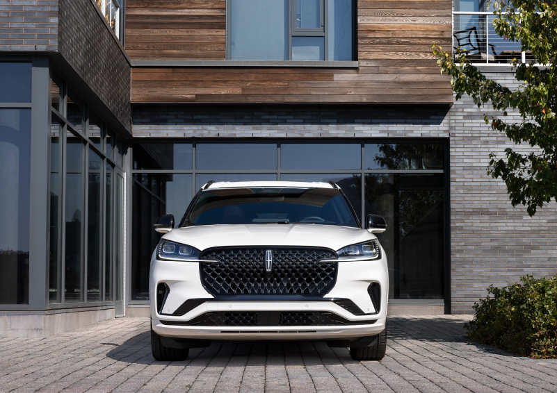 A white 2025 Lincoln Aviator® SUV is shown parked outside a modern building with gray brick and wood accents. | Mathews Lincoln in Marion OH