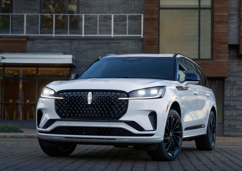 A white 2025 Lincoln Aviator® SUV is shown parked outside a modern building with gray brick and wood accents. | Mathews Lincoln in Marion OH